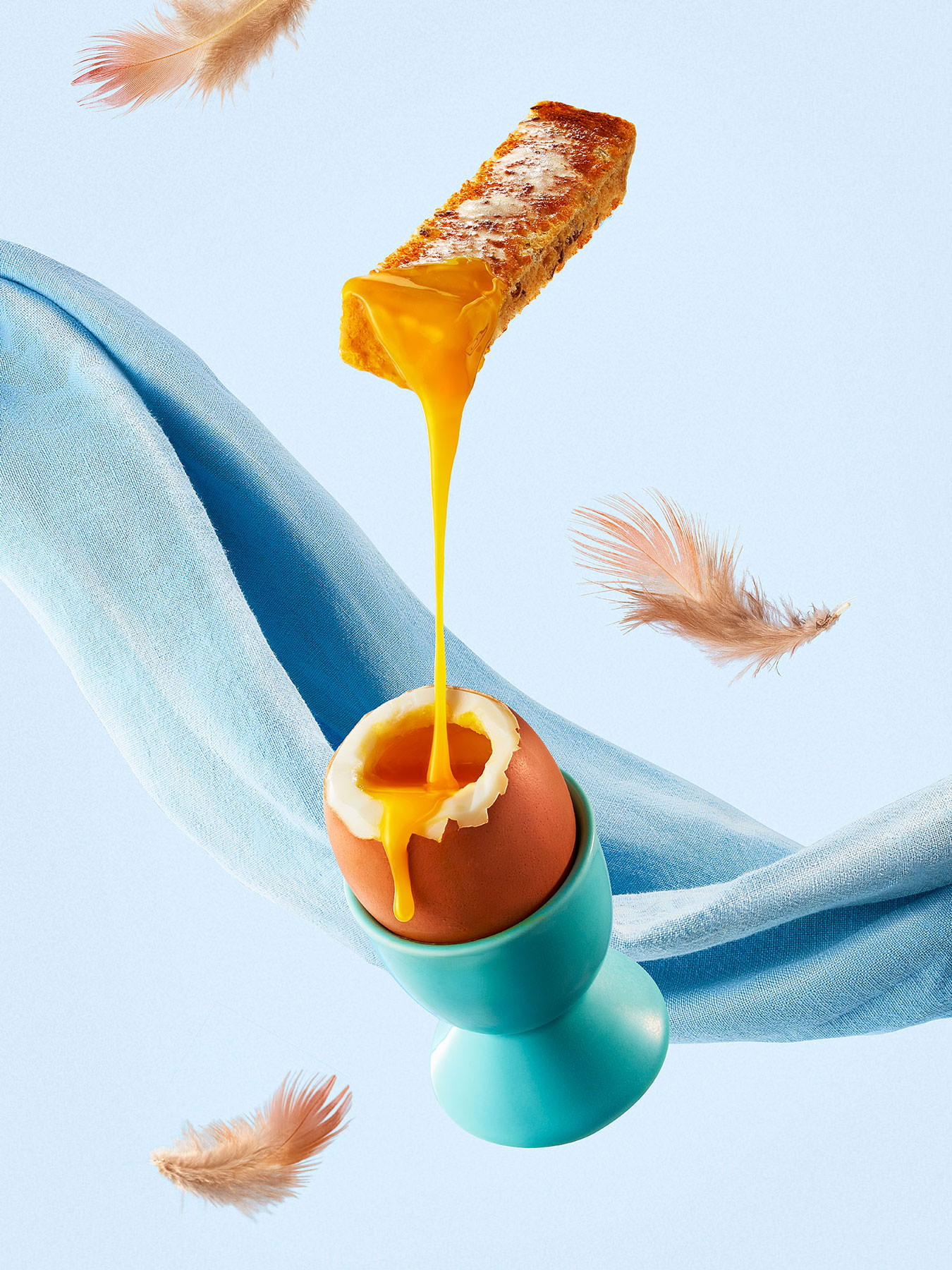 Egg and soldiers floating in mid air with runny yolk and feathers flying, set upon a blue background - London Food & Drinks Photographer