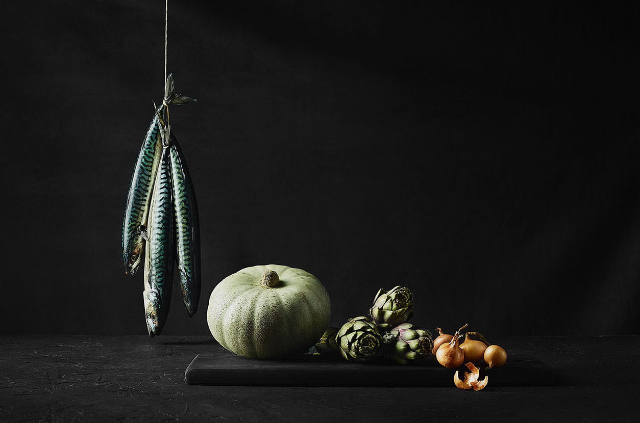 A still life photo showing a bunch of hanging mackerels, a green squash, artichokes and shallots on a black backdrop - London Food & Drinks Photographer