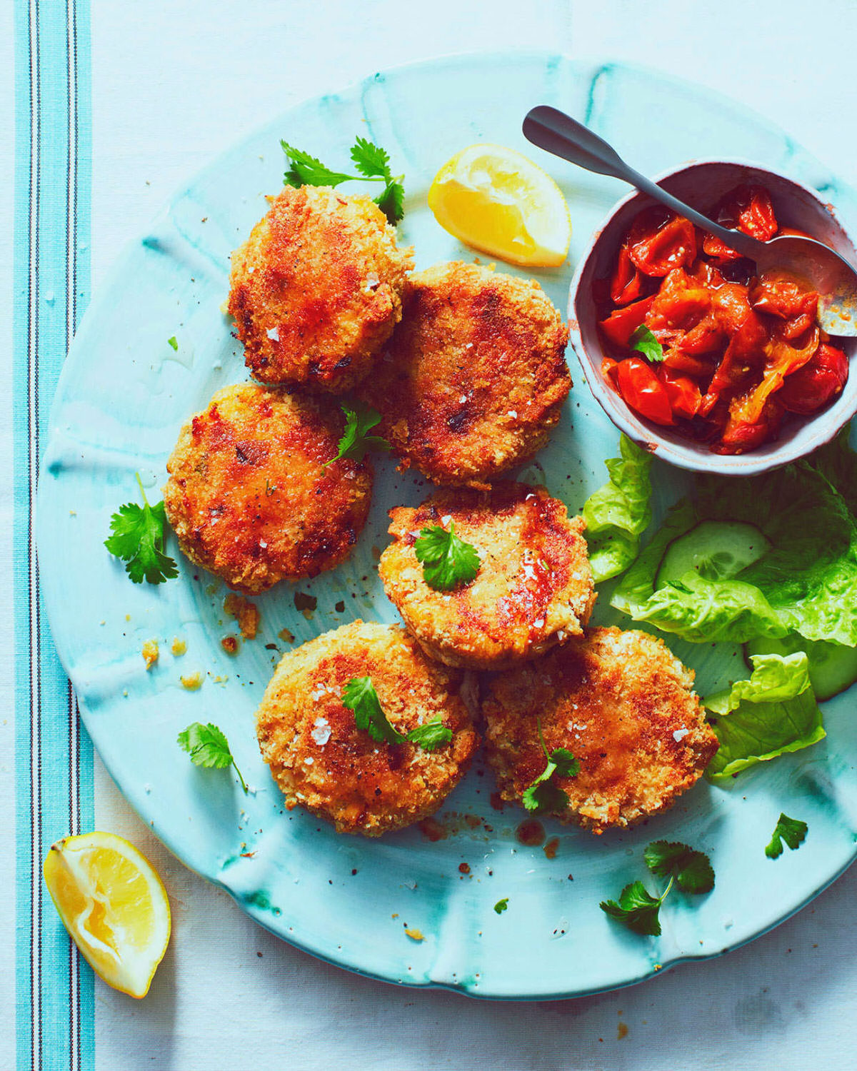 Fishcakes piled on a large turquoise platter served with a pot of fresh salsa and scattered with fresh coriander and lemon wedges